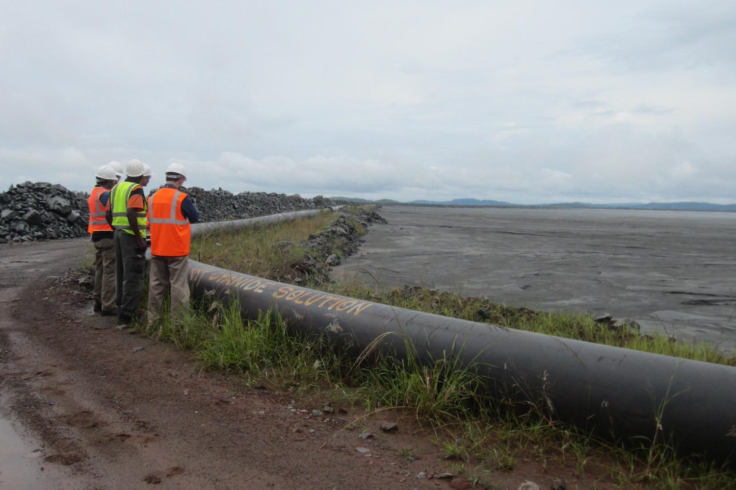 Workers stand next to a pipe labeled "cyanide solution" at a mine. 